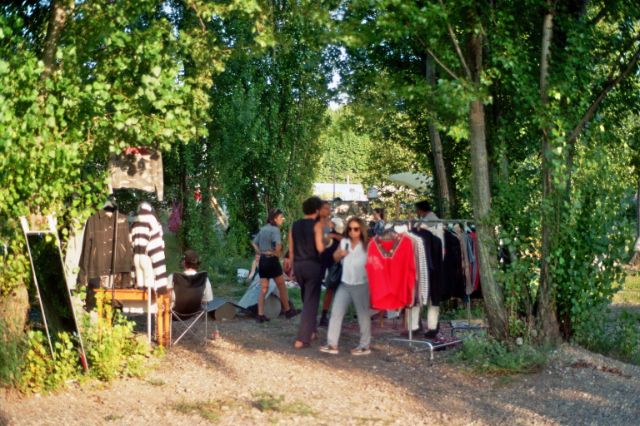 An einem Waldrand stehen Verkäufer: innen mit Kleiderbügeln und Kleidungsstücken. Sie unterhalten sich.