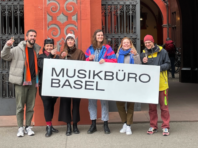 Sechs Personen halten ein Schild, auf diesem Musikbüro Basel steht. Freudige Gesichter und Prosecco in der Hand. 